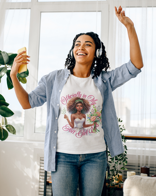 The T-shirt features the phrase "I'm in My Soft Girl Era" and depicts a graceful black woman adorned in a flowing white dress. She is holding a bouquet of vibrant flowers, each petal a burst of color against the soft fabric of her gown. The image conveys a sense of delicate beauty and the blossoming of a serene and gentle phase of life.